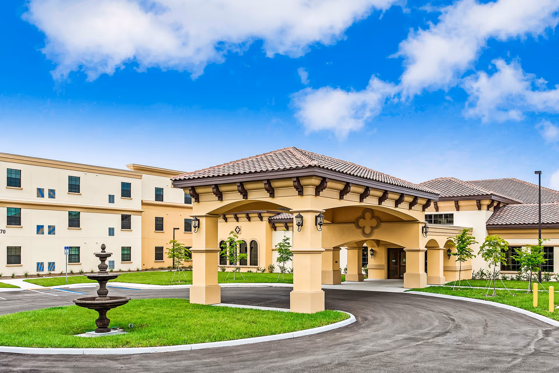 Exterior view of Discovery Commons Cypress Point showing a covered entrance with beige columns and a tiled roof, a circular driveway, green lawn with a small fountain, and a multi-story building in the background under a partly cloudy blue sky.