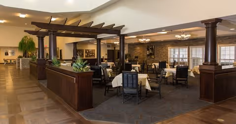 Interior view of a dining area in a senior living facility with tables covered in white tablecloths and black chairs. The space features dark wooden columns and beams, decorative plants, and warm lighting with chandeliers. Large windows allow natural light to enter the room.