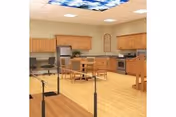 Communal activity/dining room with a kitchenette, wooden tables and chairs, hardwood floor, and parallel walking bars under a sky-patterned ceiling panel.