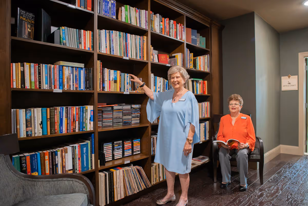 Two elderly women in a library or reading room. One woman in a light blue dress is standing and reaching for a book on a tall wooden bookshelf filled with books and CDs. The other woman, wearing an orange blouse and gray pants, is sitting on a chair holding an open book. The room has dark wooden flooring and a sign on the wall indicating 'Arts & Craft'.
