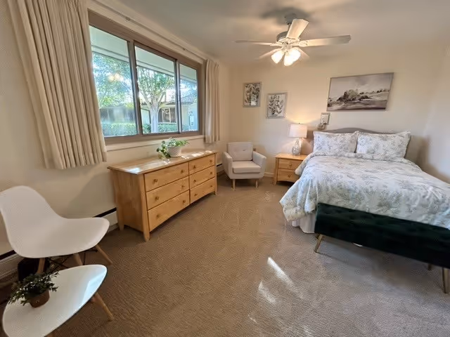 A cozy bedroom in a senior living facility with a large window showing greenery outside. The room features a bed with floral bedding, a wooden dresser, a nightstand with a lamp, a cushioned armchair, and two white chairs with a small plant on one. The walls have framed artwork, and a ceiling fan with lights is mounted above.