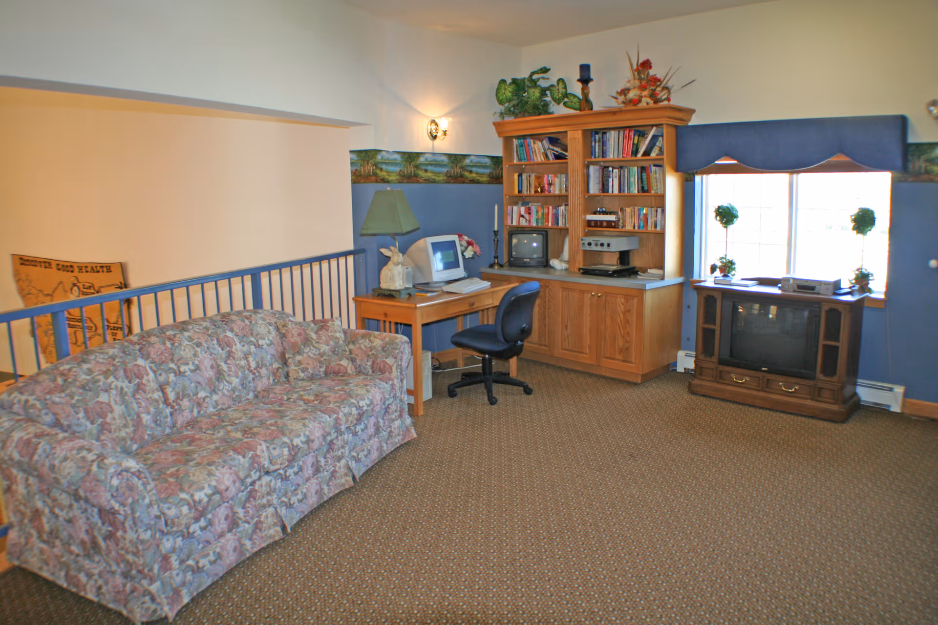 A cozy living room area with a floral patterned couch, a wooden desk with a computer and lamp, a wooden bookshelf filled with books and decorative items, and a TV on a wooden stand near a window with two small potted plants.