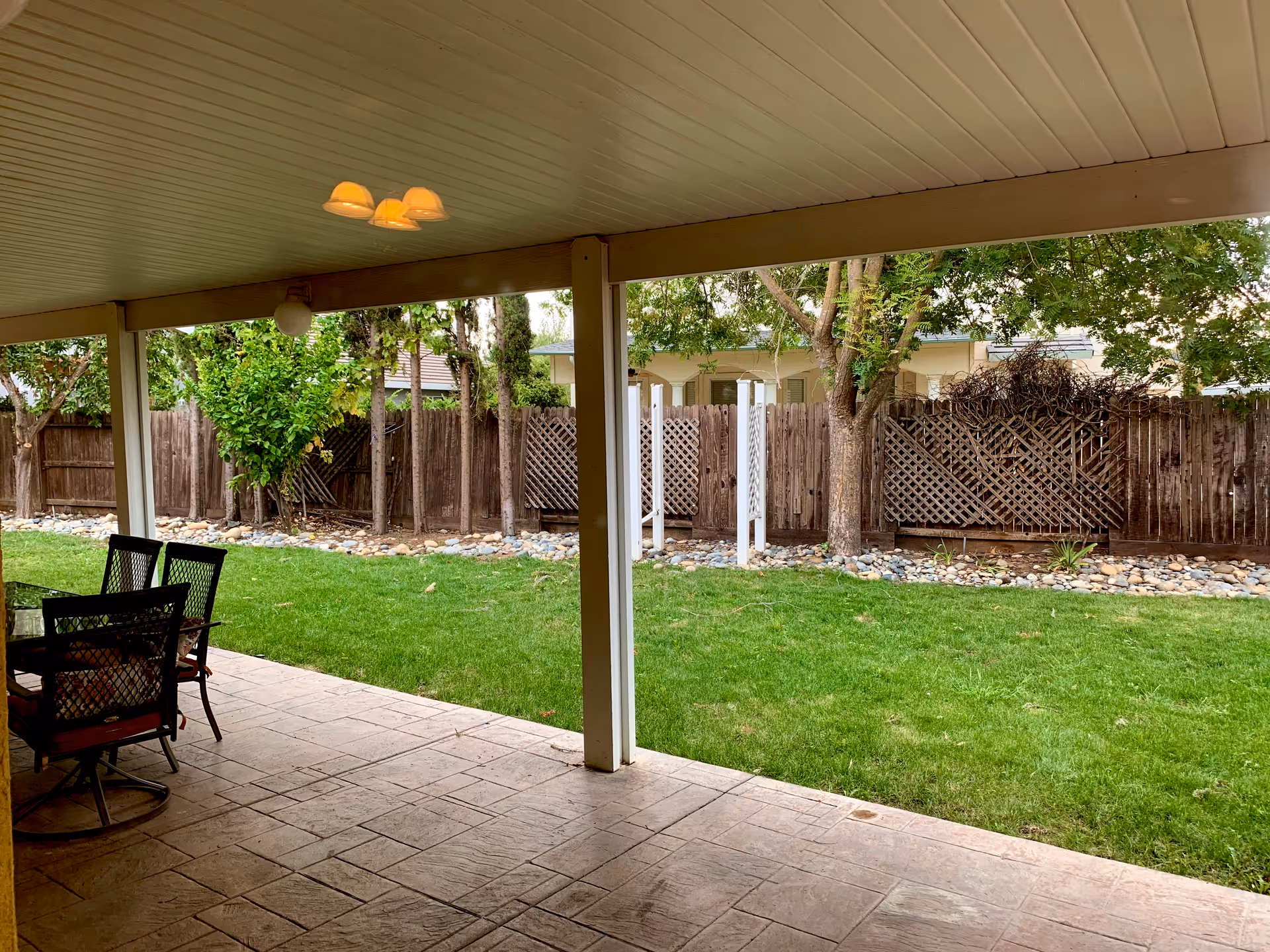 Covered patio area with a tiled floor and a table with four chairs. Beyond the patio is a green lawn bordered by a wooden fence and trees, with a white garden trellis visible in the background.