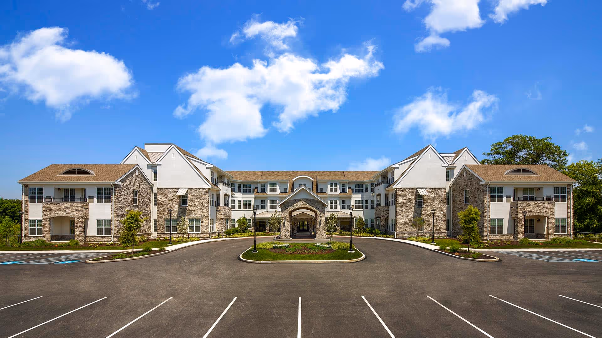 Front exterior view of a large senior living facility building with stone and white siding, multiple windows, balconies, and a covered entrance. The building is surrounded by a paved parking lot with marked parking spaces and landscaped greenery under a bright blue sky with scattered clouds.