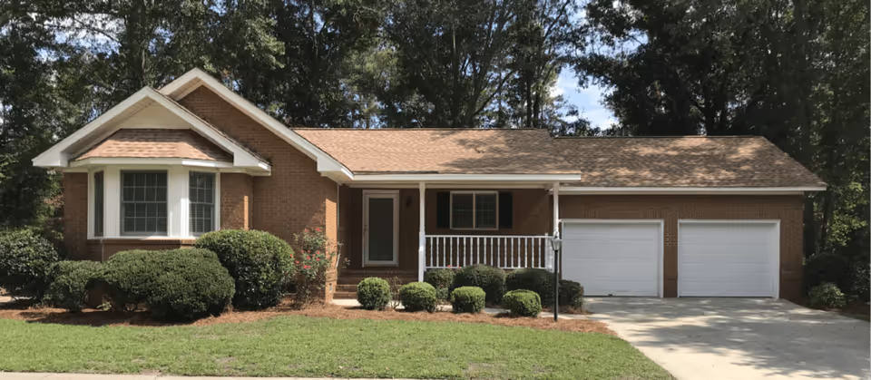 Single-story brick house with a tan shingled roof, a small front porch with white railing, a bay window on the left side, two white garage doors on the right, and neatly trimmed bushes and green lawn in front.