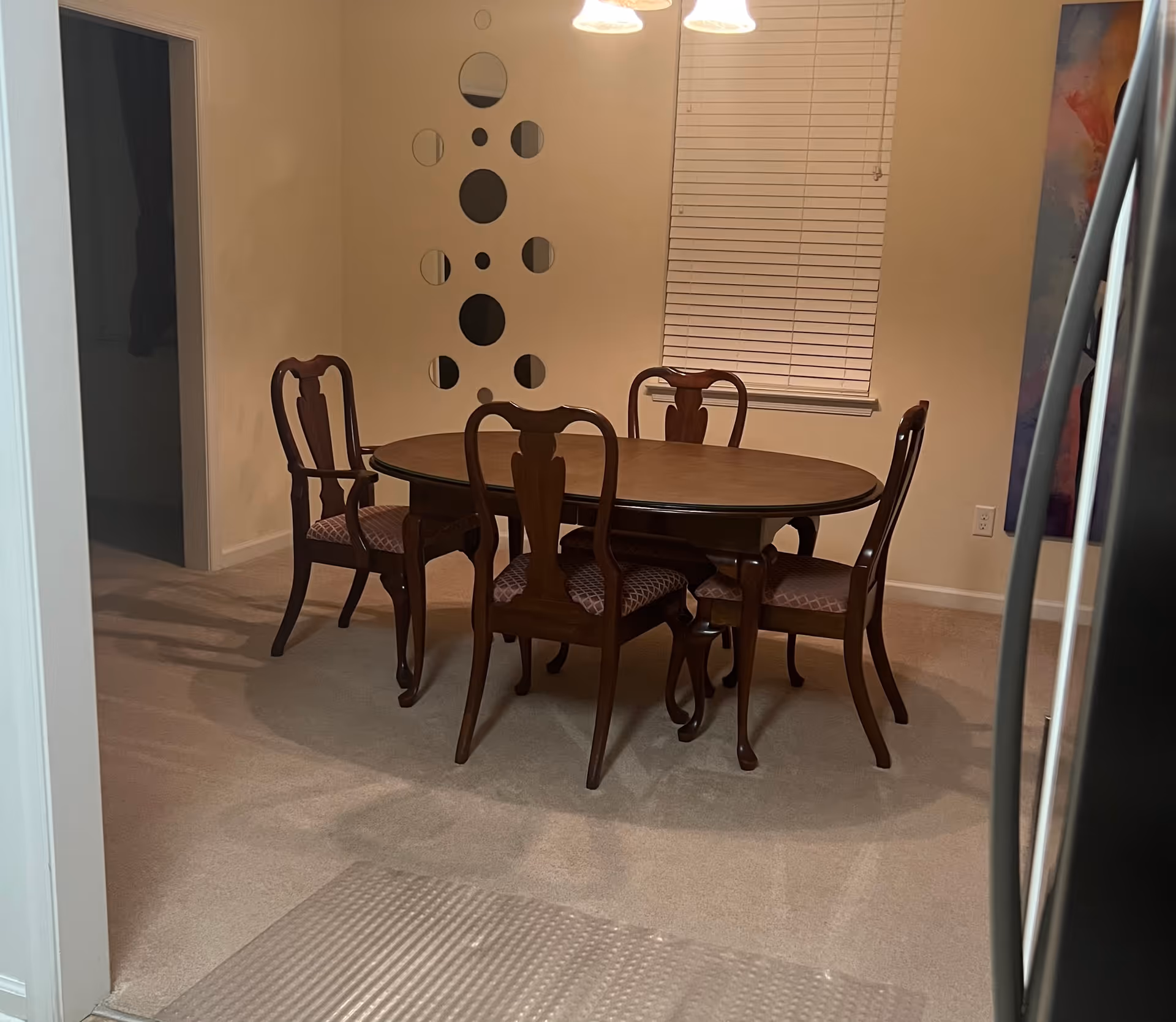 A dining area with a wooden oval table surrounded by five wooden chairs with patterned cushions. The room has beige walls, a window with closed blinds, a decorative wall mirror with circular shapes, and a colorful abstract painting partially visible on the right. The floor is carpeted, and part of a stainless steel refrigerator is visible on the right edge of the image.
