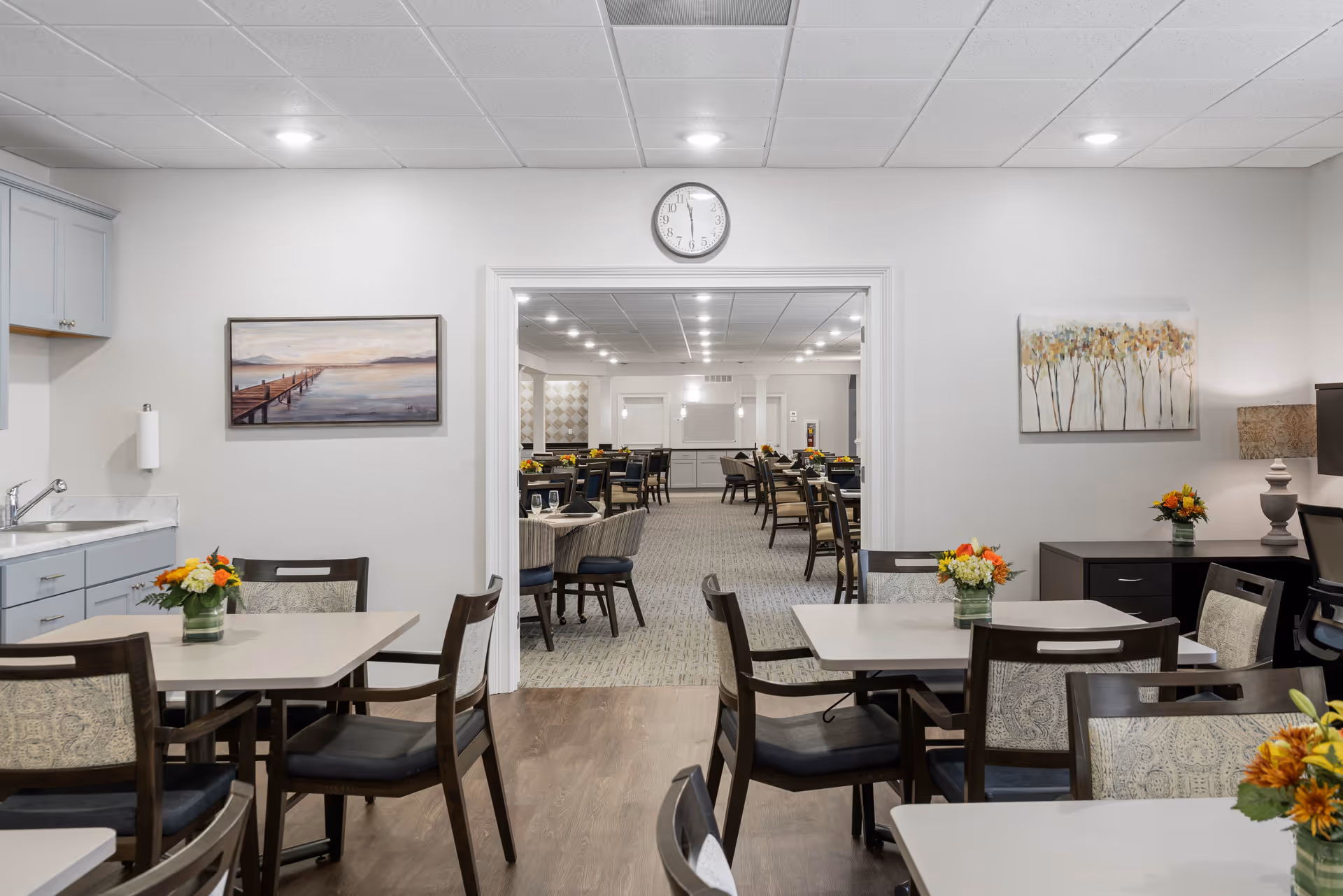 A bright and clean dining area in a senior living facility with several tables and chairs arranged neatly. Each table has a small vase with colorful flowers. The room has light-colored walls adorned with paintings, and a clock is mounted above a wide doorway that leads to a larger dining room with more tables and chairs. The ceiling has recessed lighting, and there is a small kitchenette area with a sink and cabinets on the left side.
