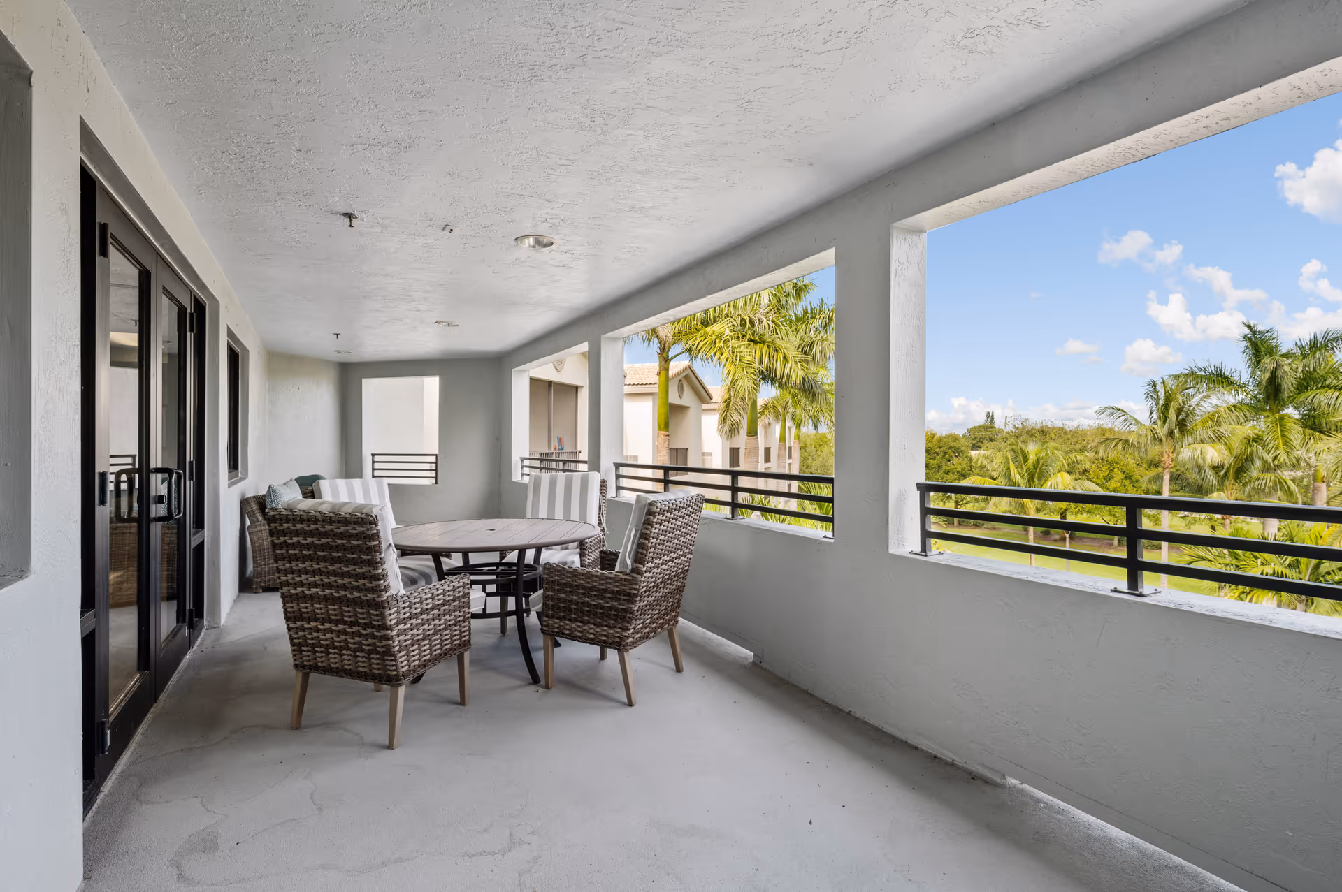 Covered outdoor balcony area with a round table and four wicker chairs with cushions. The balcony has large open windows with black railings, offering a view of palm trees and a partly cloudy sky. The walls and ceiling are light gray, and there are glass doors leading inside the building.