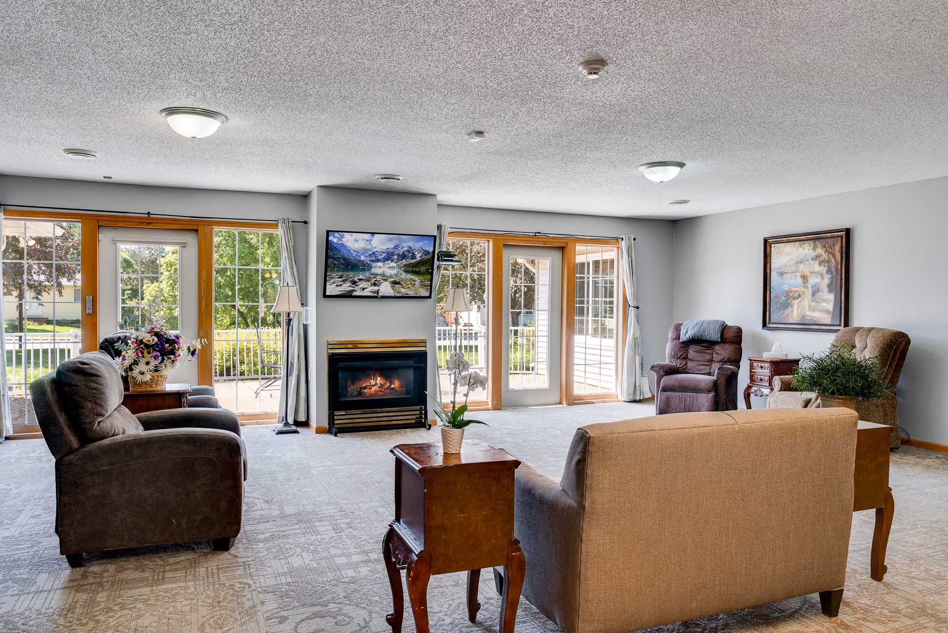 A bright and spacious living room with multiple armchairs and a sofa arranged around a central wooden table. The room features large windows and glass doors that let in natural light and offer a view of greenery outside. A fireplace with a TV mounted above it is centered on one wall, and there are decorative plants and framed artwork on the walls.