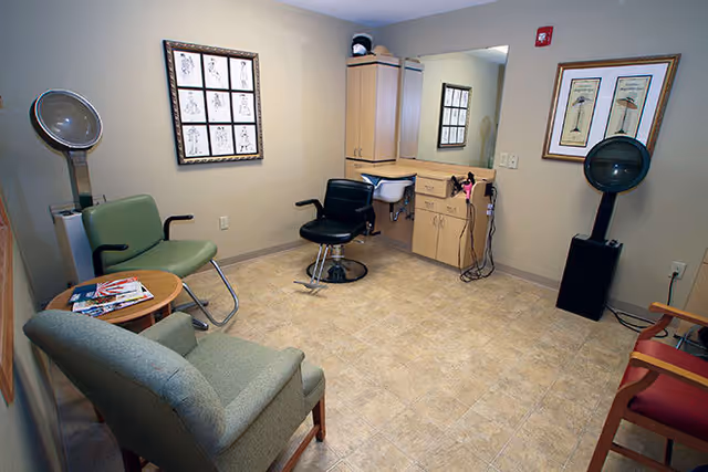 Interior view of a small salon or hair care room with a black salon chair in front of a mirror and cabinet, a green chair, a gray armchair, a red chair, a round wooden table with magazines, and two hair dryers mounted on stands against the walls.
