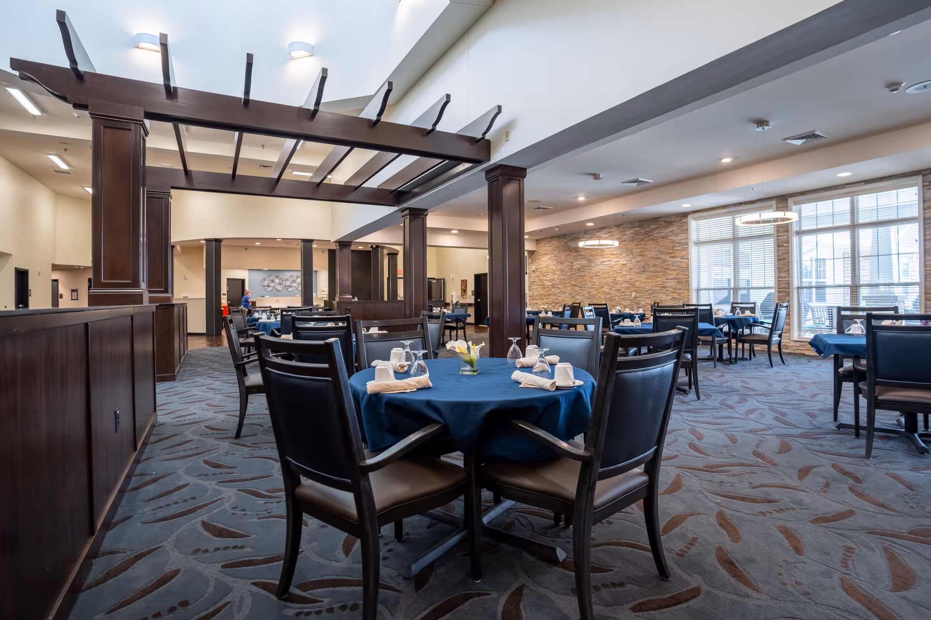 A spacious dining room with multiple round tables covered with blue tablecloths, each set with napkins, glasses, and small flower centerpieces. The room features dark wooden pillars and beams, a patterned carpet, large windows letting in natural light, and modern circular light fixtures on the ceiling.