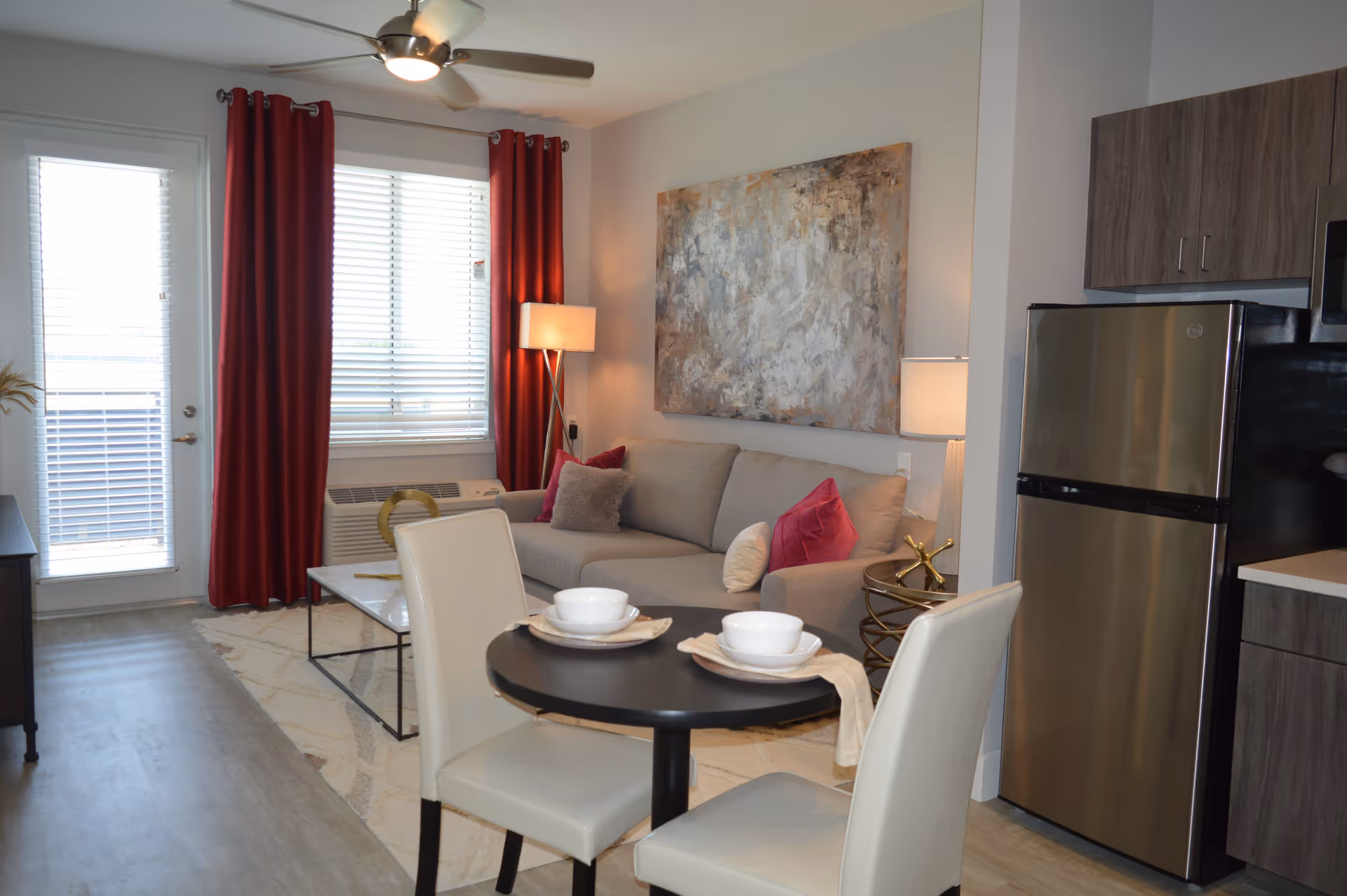 A modern senior living apartment interior featuring a small round dining table set for two with white chairs and bowls, a gray sofa with colorful pillows, a large abstract painting on the wall, a floor lamp, red curtains covering a window and door, and a stainless steel refrigerator next to wooden kitchen cabinets.