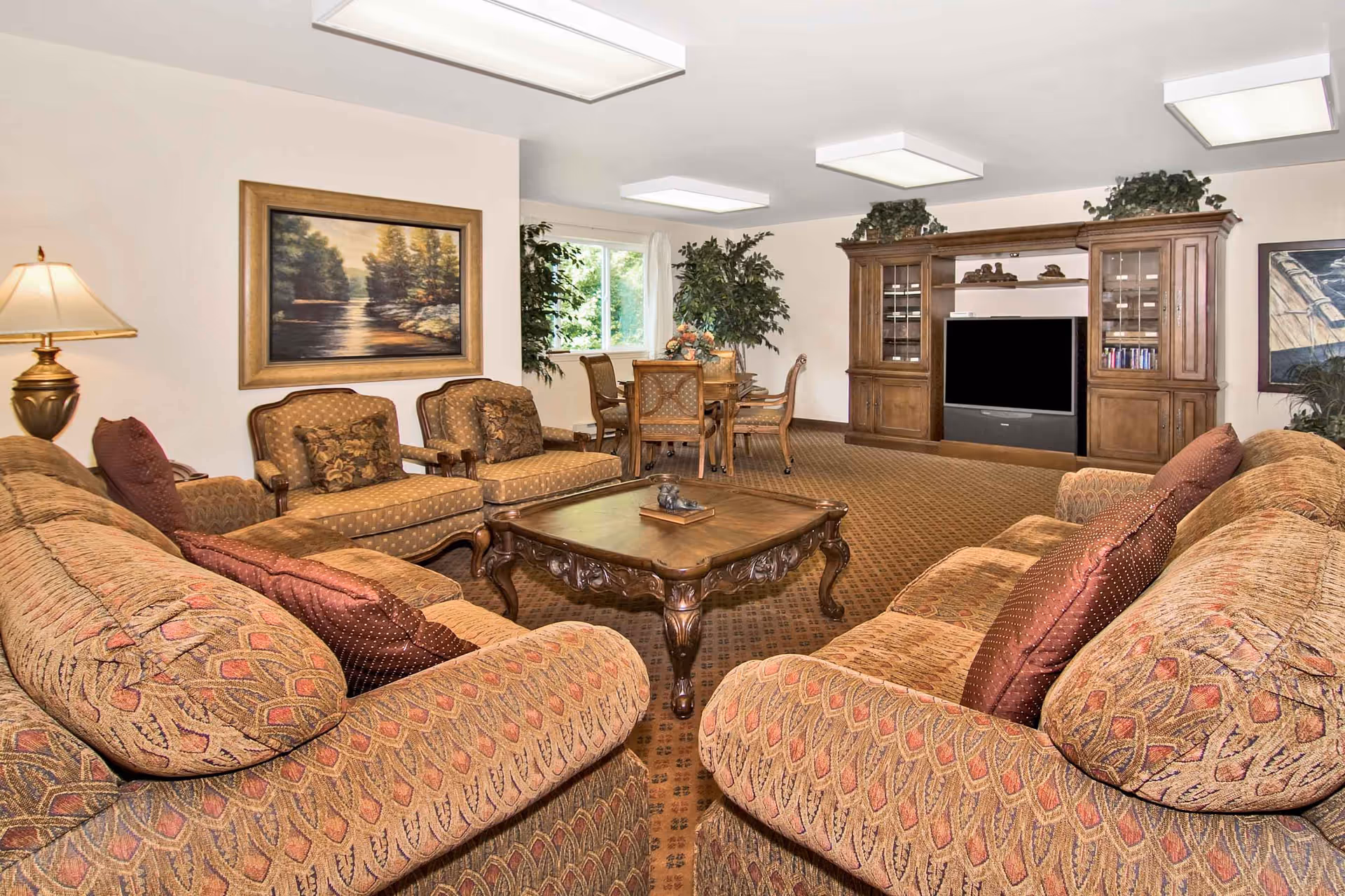 Spacious furnished living room with patterned sofas and armchairs arranged around a wooden coffee table, TV cabinet, dining table, and wall art.
