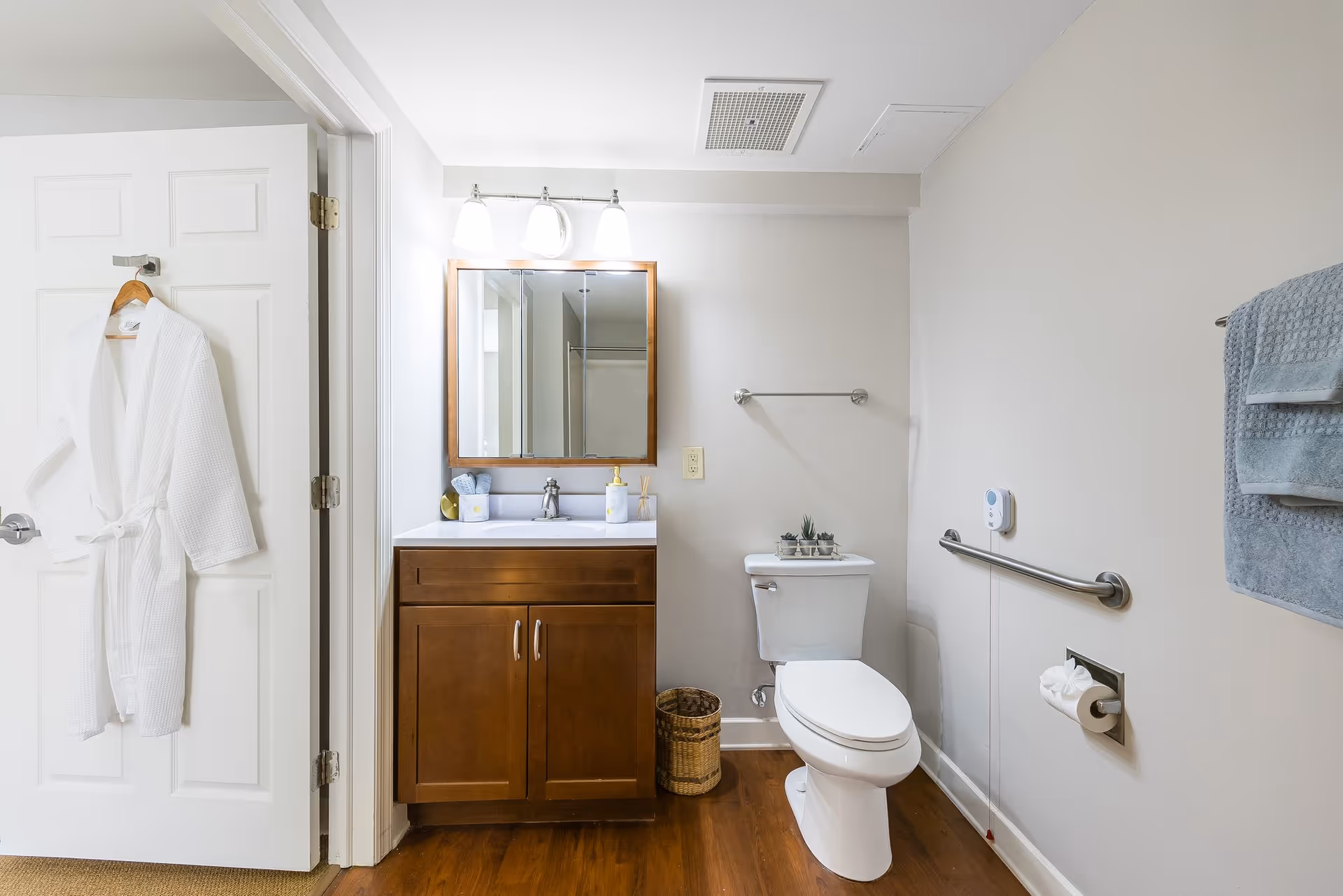 A bright bathroom with a wooden vanity and mirror, a toilet with a grab bar, towel racks, and a robe hanging on the door.