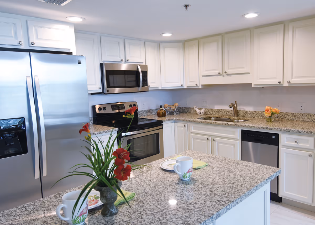 Bright modern kitchen with stainless steel appliances, white cabinets, granite countertops and an island topped with flowers and mugs.