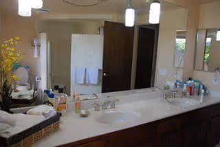 A bathroom interior featuring a double sink vanity with various toiletries on the countertop, a large mirror above the sinks, a towel rack with white towels, and a door leading to another room. The lighting includes hanging pendant lights and a wall-mounted fixture.