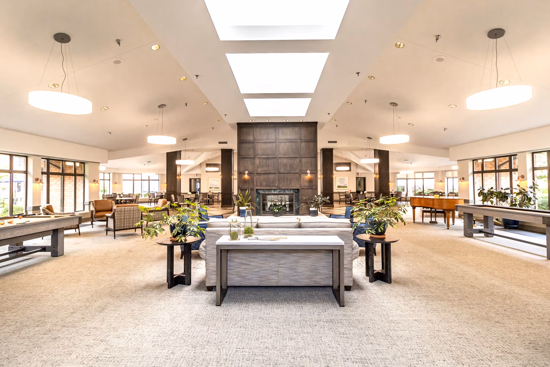 Spacious bright senior living common lounge with sofas, plants, a central fireplace, skylights, and game tables.