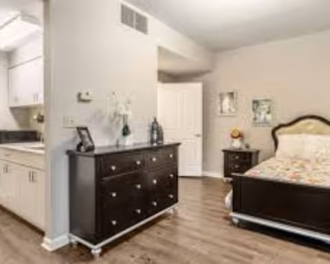 Interior view of a bedroom with a dark wooden bed frame and matching dresser. The bed has a colorful quilt, and there is a nightstand with a lamp and decorative items. The room has light-colored walls and wood flooring. A doorway leads to another room, and part of a kitchen area with white cabinets is visible on the left side.