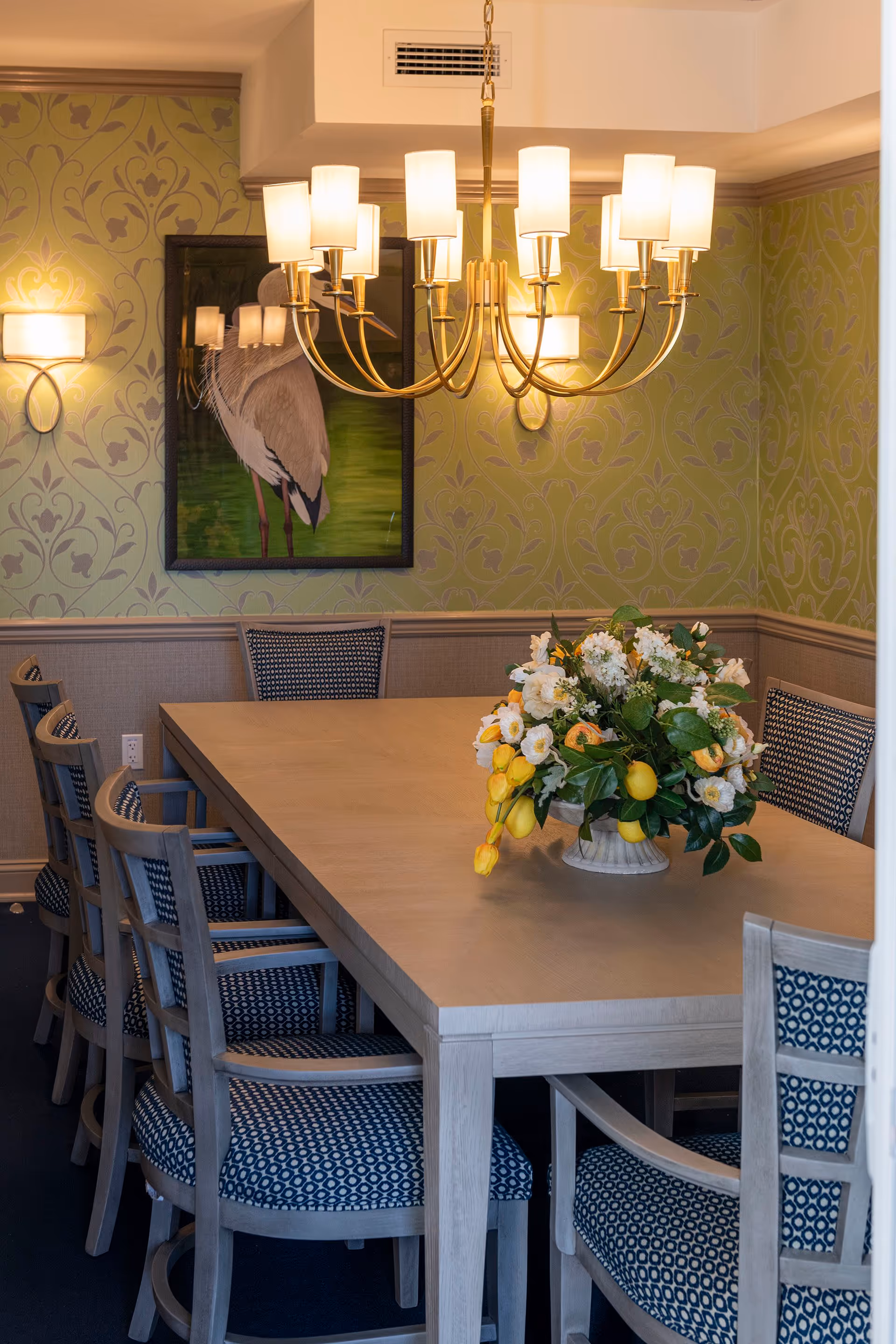 Well-lit dining room with a long wooden table surrounded by patterned chairs, a floral centerpiece, chandelier, and green patterned wallpaper.