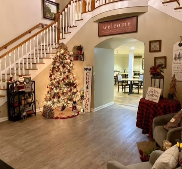 Interior view of a senior living facility decorated for Christmas with a lit Christmas tree, holiday decorations including a 'Merry Christmas' sign, and a 'welcome' sign above an archway leading to a dining area with tables and chairs.
