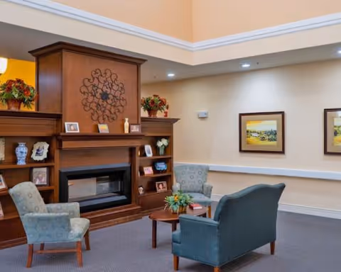 A cozy senior living common area with a wooden fireplace mantel decorated with framed photos, vases, and flower arrangements. There are two armchairs and a loveseat arranged around a small wooden coffee table with a plant on it. The walls are light-colored with two framed landscape paintings, and the room has recessed lighting and a high ceiling.