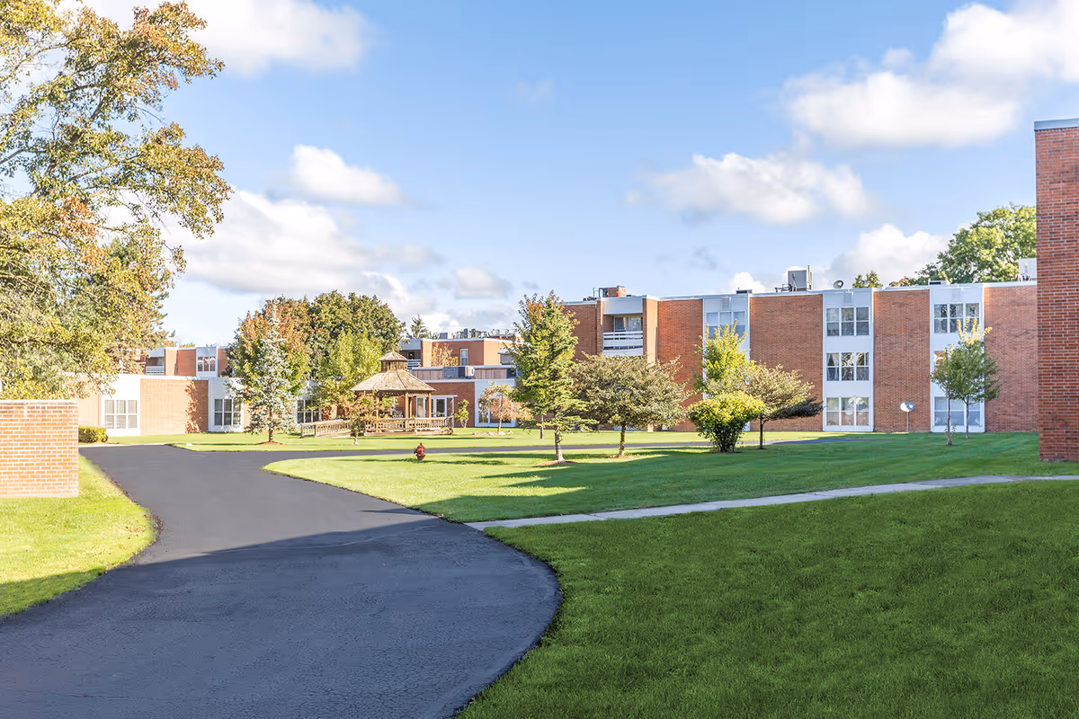 Outdoor view of a senior living facility with a paved pathway, green lawn, several trees, and a wooden gazebo. The building is made of red brick with multiple windows and a clear blue sky with some clouds above.