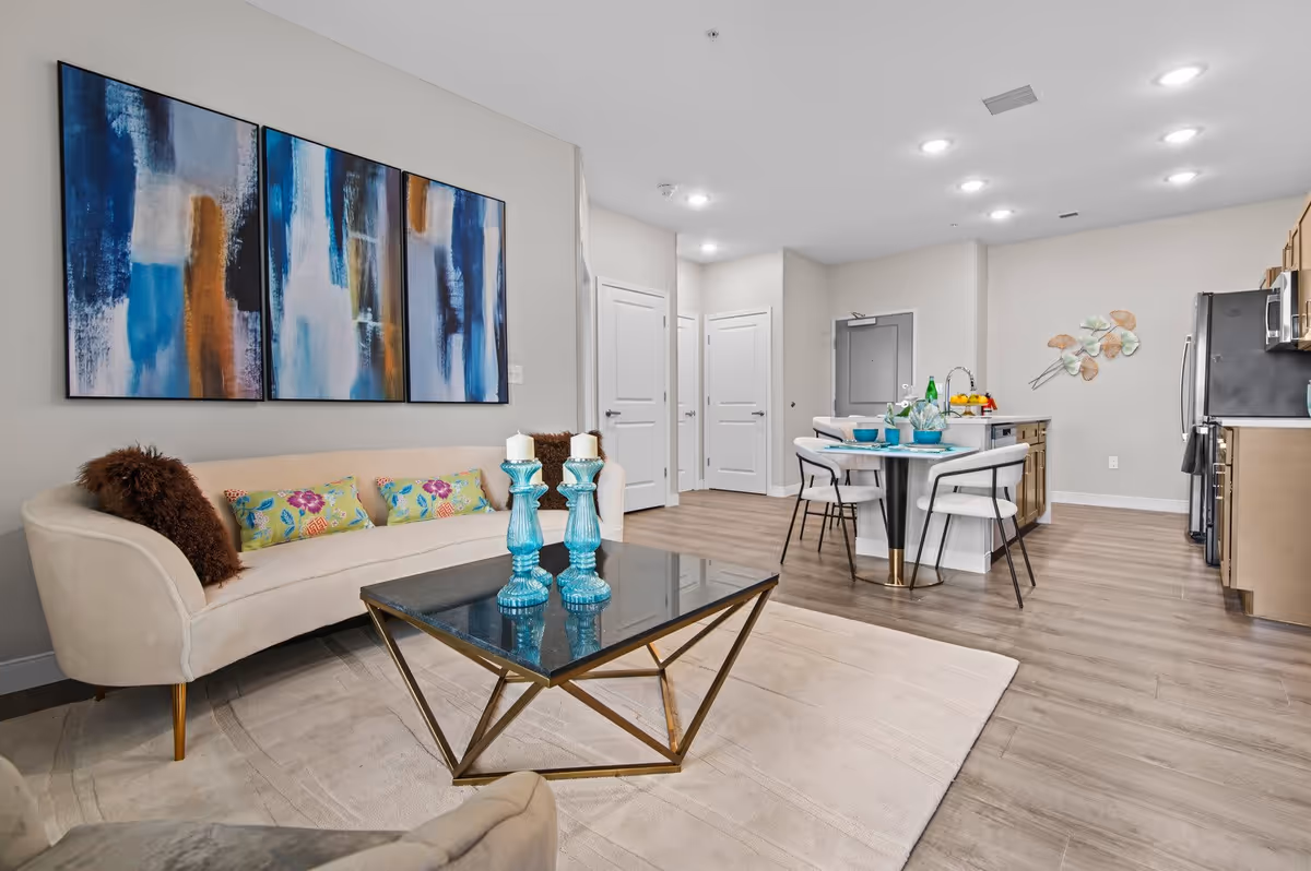 Modern open-concept living area featuring a beige curved sofa with colorful pillows, a glass coffee table with blue candle holders, abstract blue and brown artwork on the wall, and a kitchen with a dining table set for four with blue dishes and light-colored chairs. The space has light wood flooring and recessed ceiling lights.
