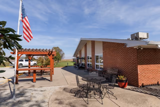 Outdoor patio area at Falls City Nursing and Rehabilitation Center featuring a brick building with large windows, metal tables and chairs, a wooden pergola with benches, an American flag on a flagpole, and a clear blue sky.