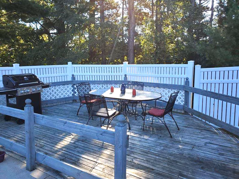 Outdoor wooden deck area with a round table and four metal chairs with red cushions. There are three condiment bottles on the table. A black grill is positioned on the left side of the deck. The deck is enclosed by a white fence with lattice panels and surrounded by trees in the background.
