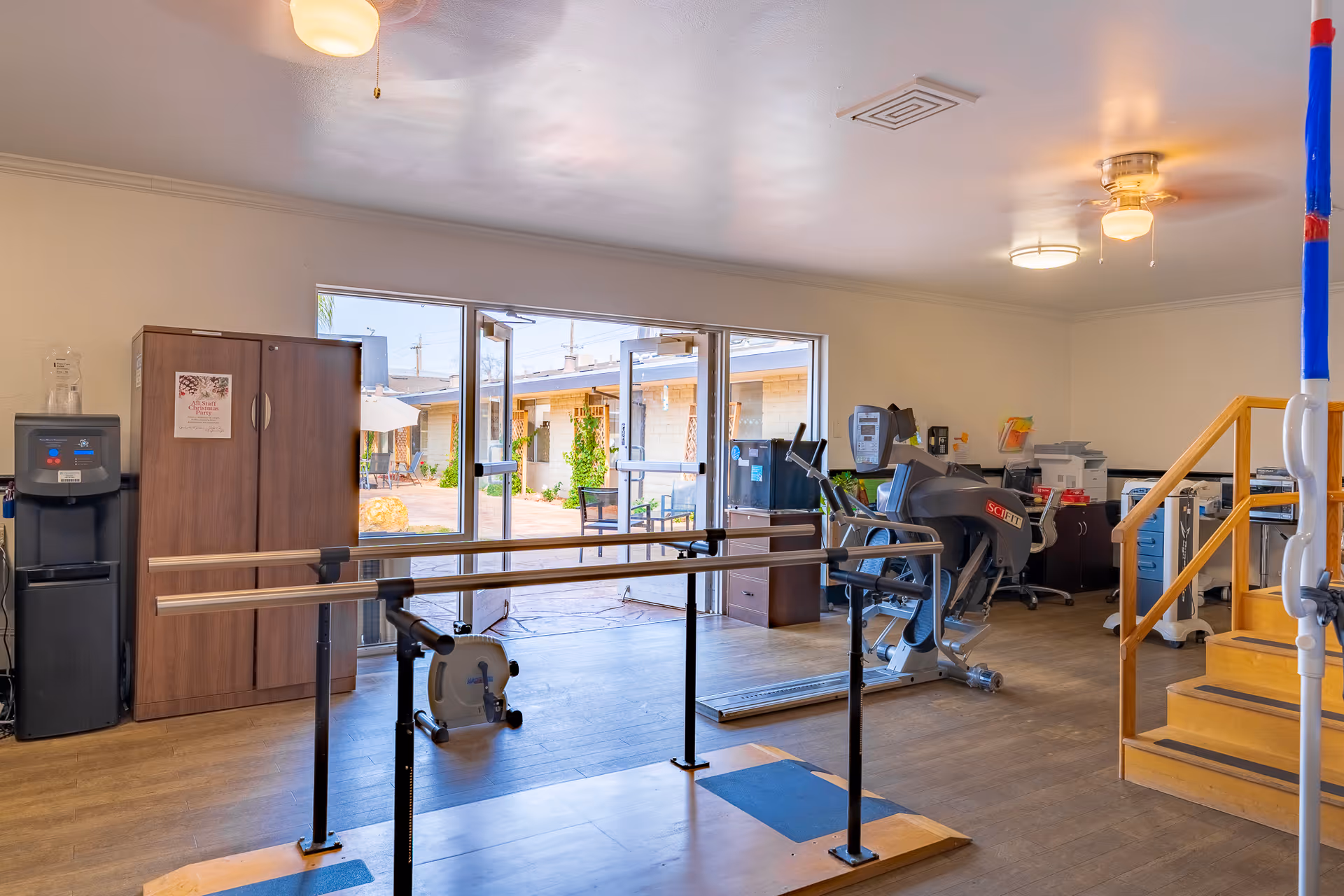 Rehabilitation/therapy room with parallel bars, exercise equipment, a stationary bike, and open doors to an outdoor courtyard.