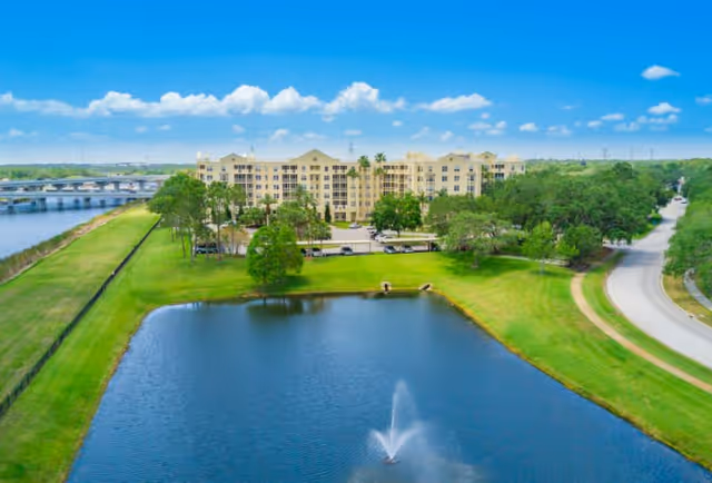 Aerial view of Stratford Court of Palm Harbor, a large multi-story residential building surrounded by green lawns, trees, a pond with a fountain, and a nearby road and bridge under a blue sky with scattered clouds.