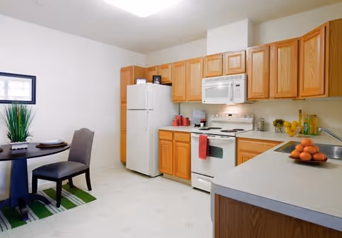 Bright kitchen with oak cabinets, white appliances, a small dining table and a counter with fruit.