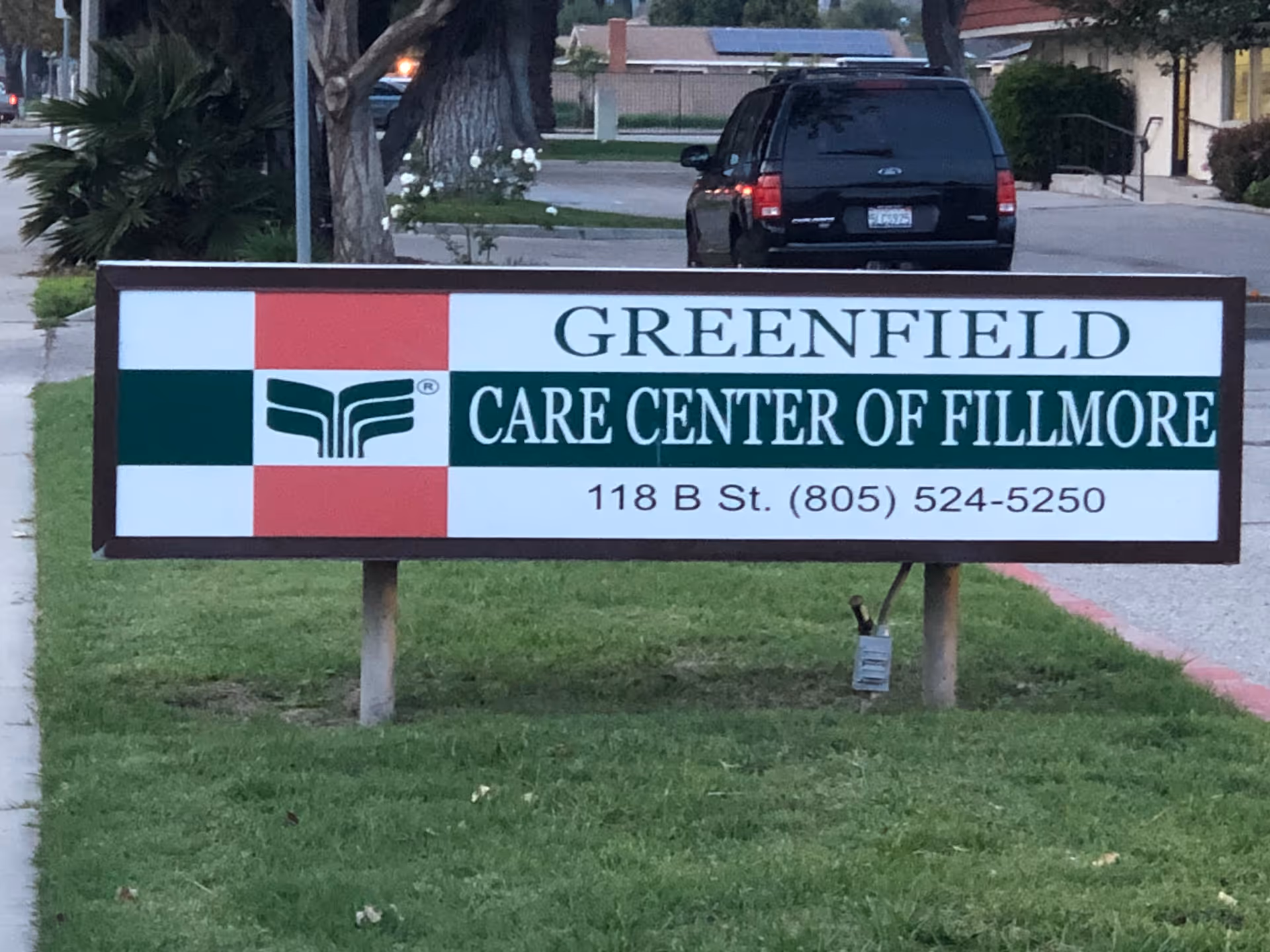A roadside sign reading "Greenfield Care Center of Fillmore" on a grassy lawn with a parked SUV and sidewalk behind it.