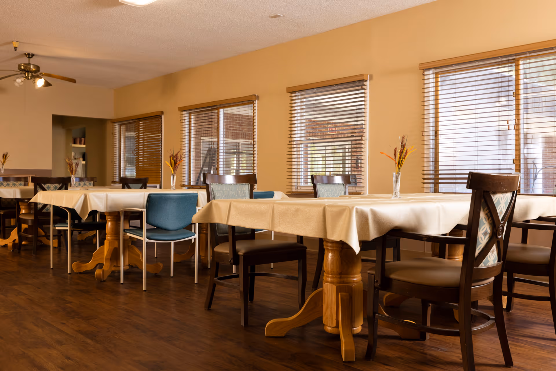 Bright dining room with multiple tables covered in tablecloths, chairs, and small vases of flowers by windows.