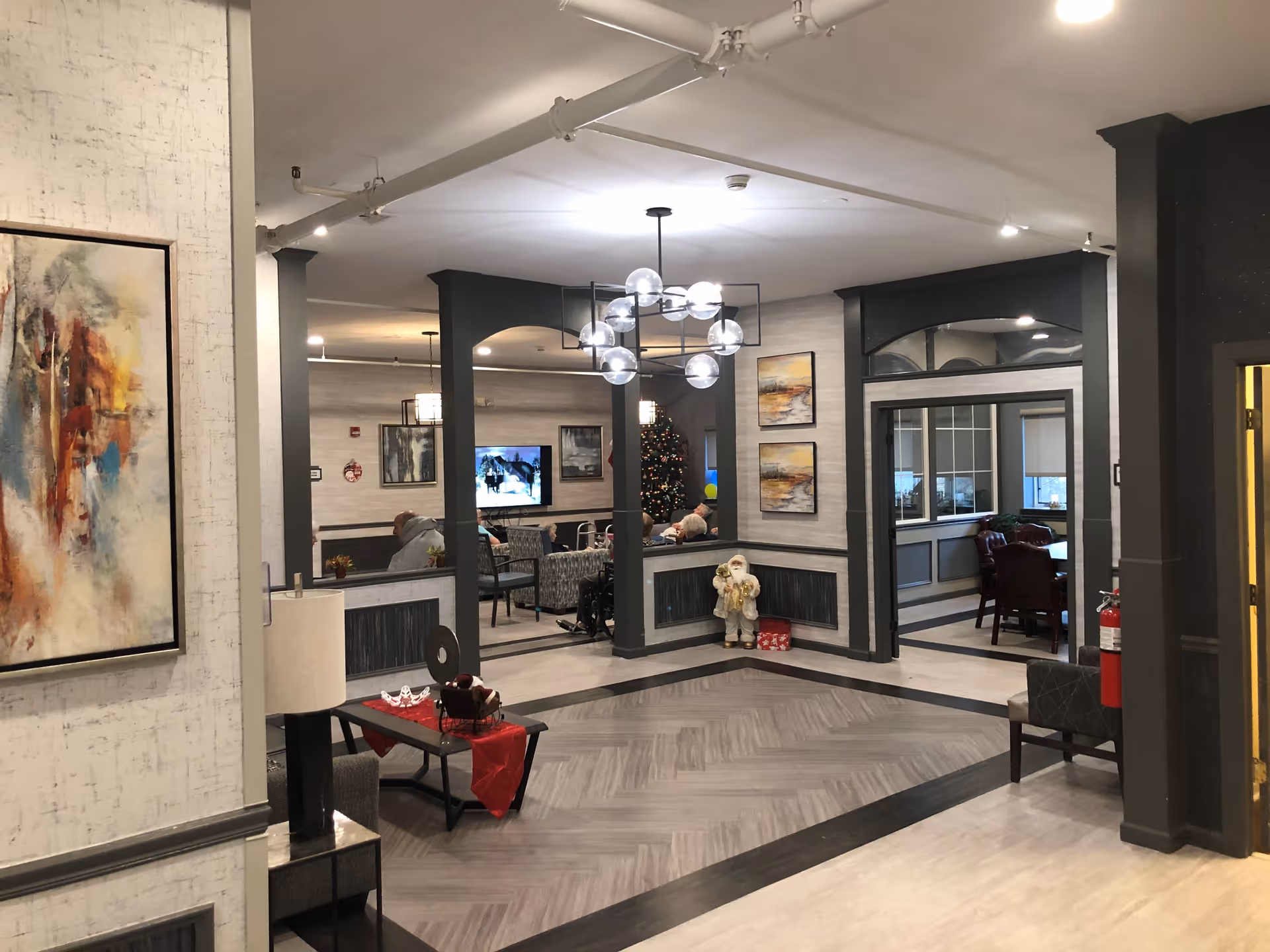 Interior view of a senior living facility common area with modern decor. The space features a seating area with chairs and a television, a Christmas tree, abstract paintings on the walls, a decorative Santa figure, and a contemporary chandelier. There is a small table with a red runner and a lamp, and a fire extinguisher mounted on the wall near a doorway.