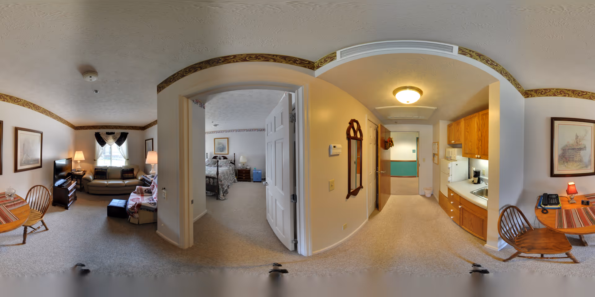 A panoramic view of a senior living apartment at The Neighborhood of Somerset showing a living room with a sofa, TV, and chairs, a bedroom with a bed and nightstands, and a kitchenette with wooden cabinets, a sink, and appliances. The space is decorated with framed pictures, a mirror, and lamps, with carpeted floors and neutral-colored walls.