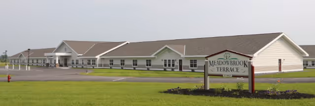 Long single-story assisted living building with a Meadowbrook Terrace sign on a grassy lawn and parking lot.