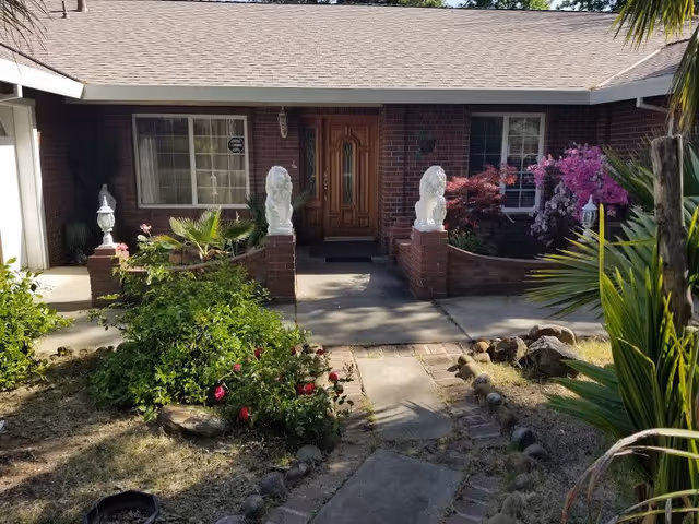 Front entrance of a single-story brick building with a wooden door flanked by two white lion statues on brick pillars. There are windows on either side of the door, and the entrance is surrounded by various green plants and colorful flowers. A concrete pathway leads up to the door.