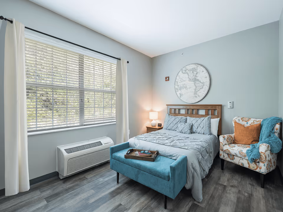 A bright bedroom with a large window covered by white curtains and blinds. The room features a wooden bed with a gray comforter and pillows, a small wooden nightstand with a lamp, and a decorative round map hanging above the bed. In front of the bed is a teal upholstered bench with a tray on top. To the right is a patterned armchair with an orange pillow and a teal throw blanket. The floor is covered with wood-style flooring.