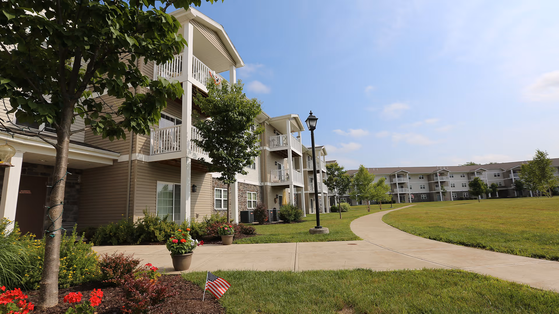 Exterior view of Schuyler Commons senior living facility showing a multi-story building with balconies, a paved walkway, green lawn, trees, flower pots, and a clear blue sky.