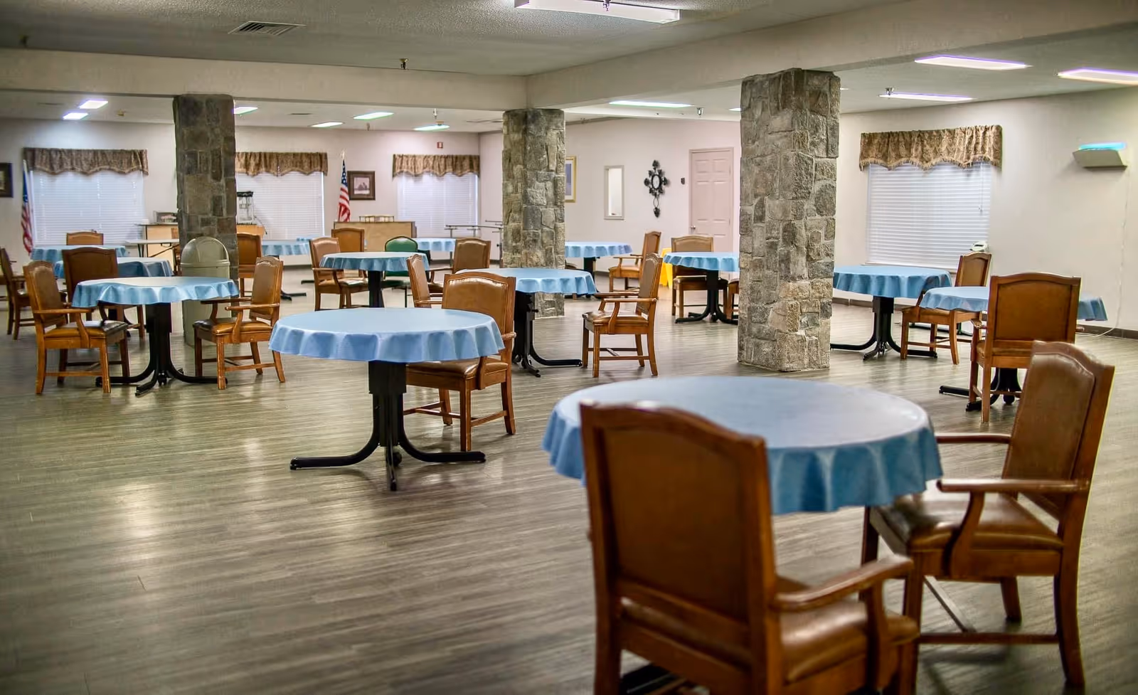 A spacious dining room with multiple round tables covered with light blue tablecloths, each surrounded by wooden chairs with armrests. The room features stone pillars, large windows with valances, and a wooden floor. The lighting is bright and the room appears clean and orderly.