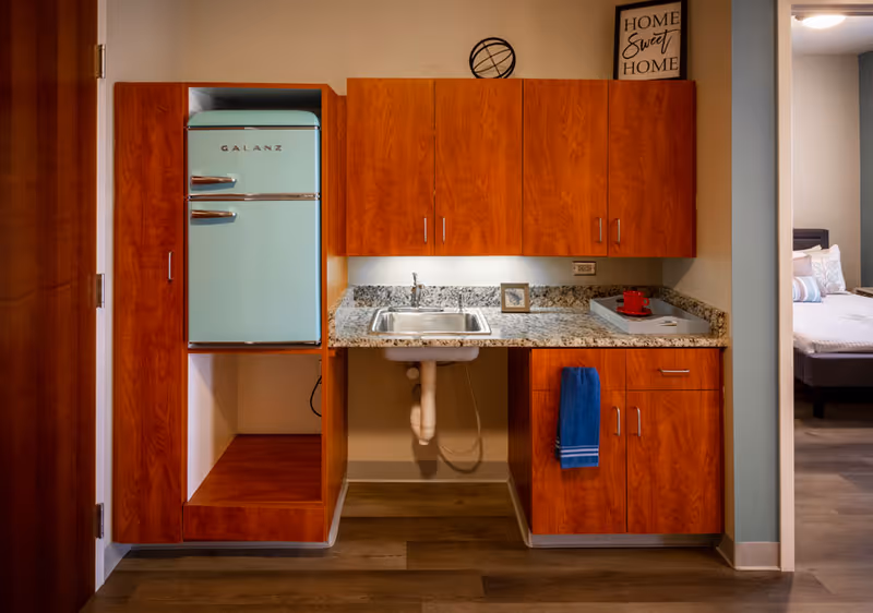 A small kitchenette area with wooden cabinets, a granite countertop, and a stainless steel sink. A retro-style mint green Galanz refrigerator is built into the cabinetry on the left. A blue towel hangs from a cabinet handle below the sink. On the countertop, there is a gray tray with a red cup and saucer. A framed picture and a decorative black wire sphere are placed on top of the cabinets. To the right, a doorway leads to a bedroom with a bed and pillows visible.