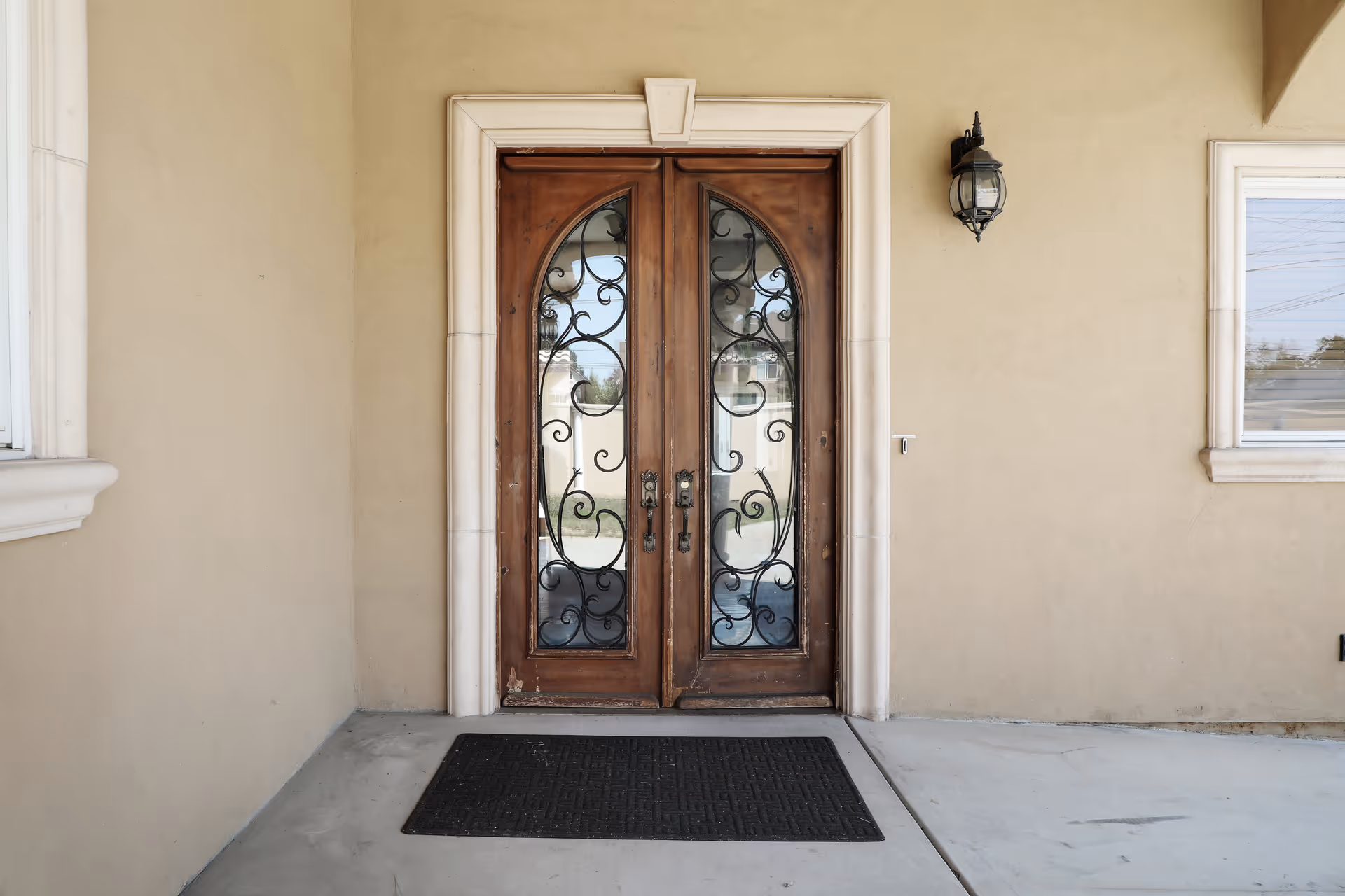 Double wooden front doors with decorative wrought iron glass panels, set in a beige stucco wall with a black outdoor wall lantern on the right side and a black doormat on the concrete floor in front of the doors.