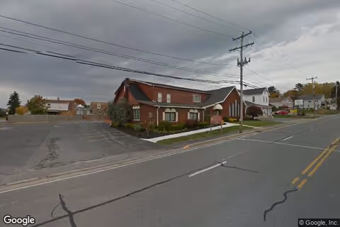 Street view of a brick building with a sloped roof and adjacent white houses along a road under a cloudy sky. There are power lines and a parking area next to the building.