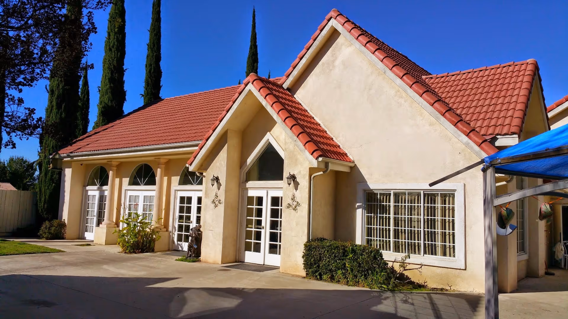 Exterior view of a single-story building with beige stucco walls and red tile roofing under a clear blue sky. The building has multiple white-framed glass doors and windows, with some greenery and tall cypress trees nearby. A blue canopy is partially visible on the right side.
