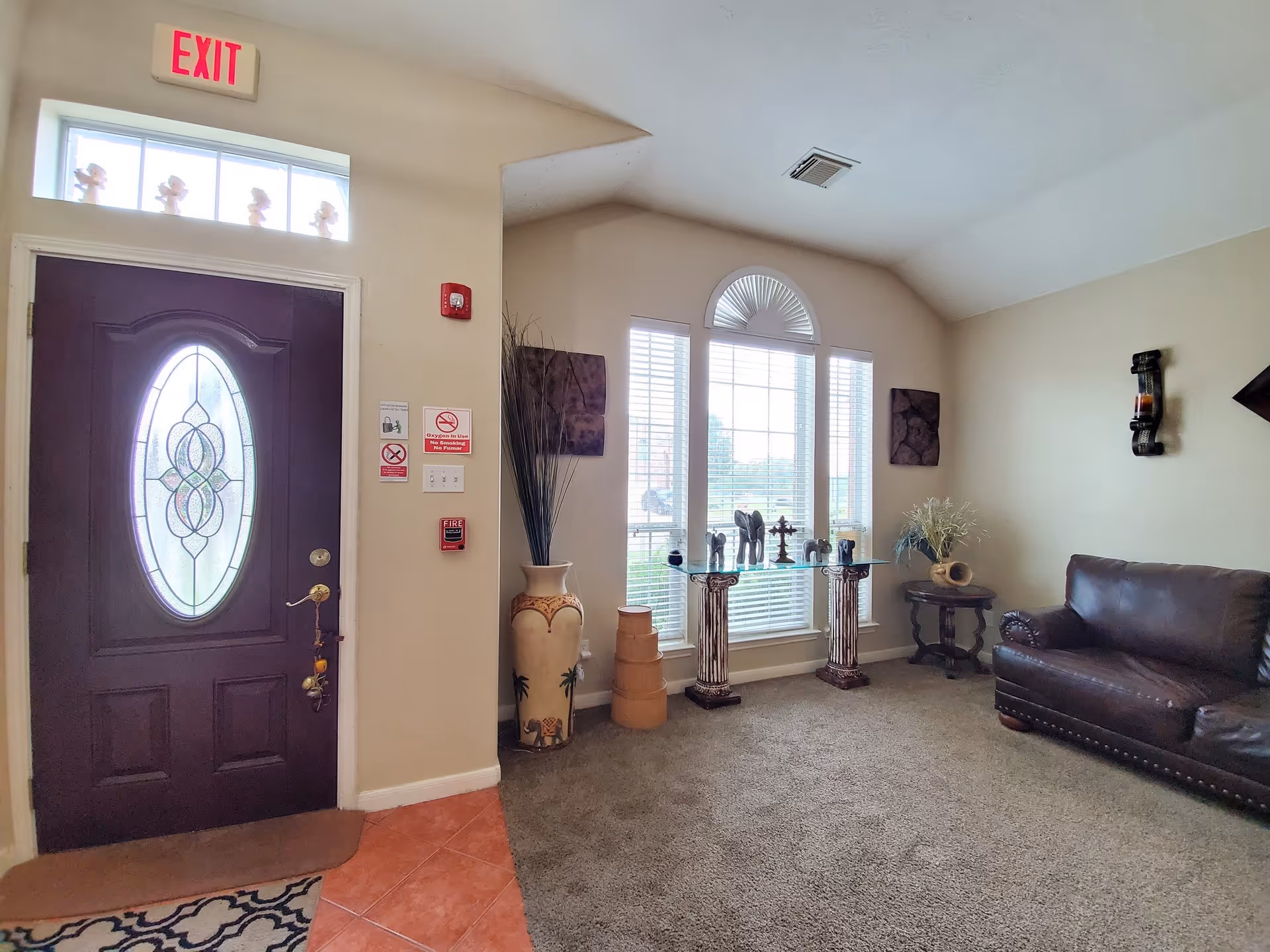 Interior view of a living room area in an assisted living facility featuring a dark wooden front door with decorative glass, an exit sign above it, a large window with white blinds and an arched top, a glass table with decorative items including small elephant statues and a cross, a tall vase with decorative branches, a small round side table with a plant, and a dark brown leather couch on a carpeted floor.