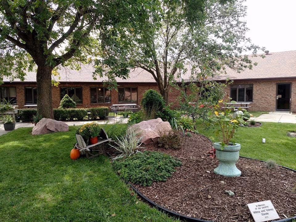 Courtyard garden with trees, potted plants, rocks and benches in front of a single-story brick retirement facility.