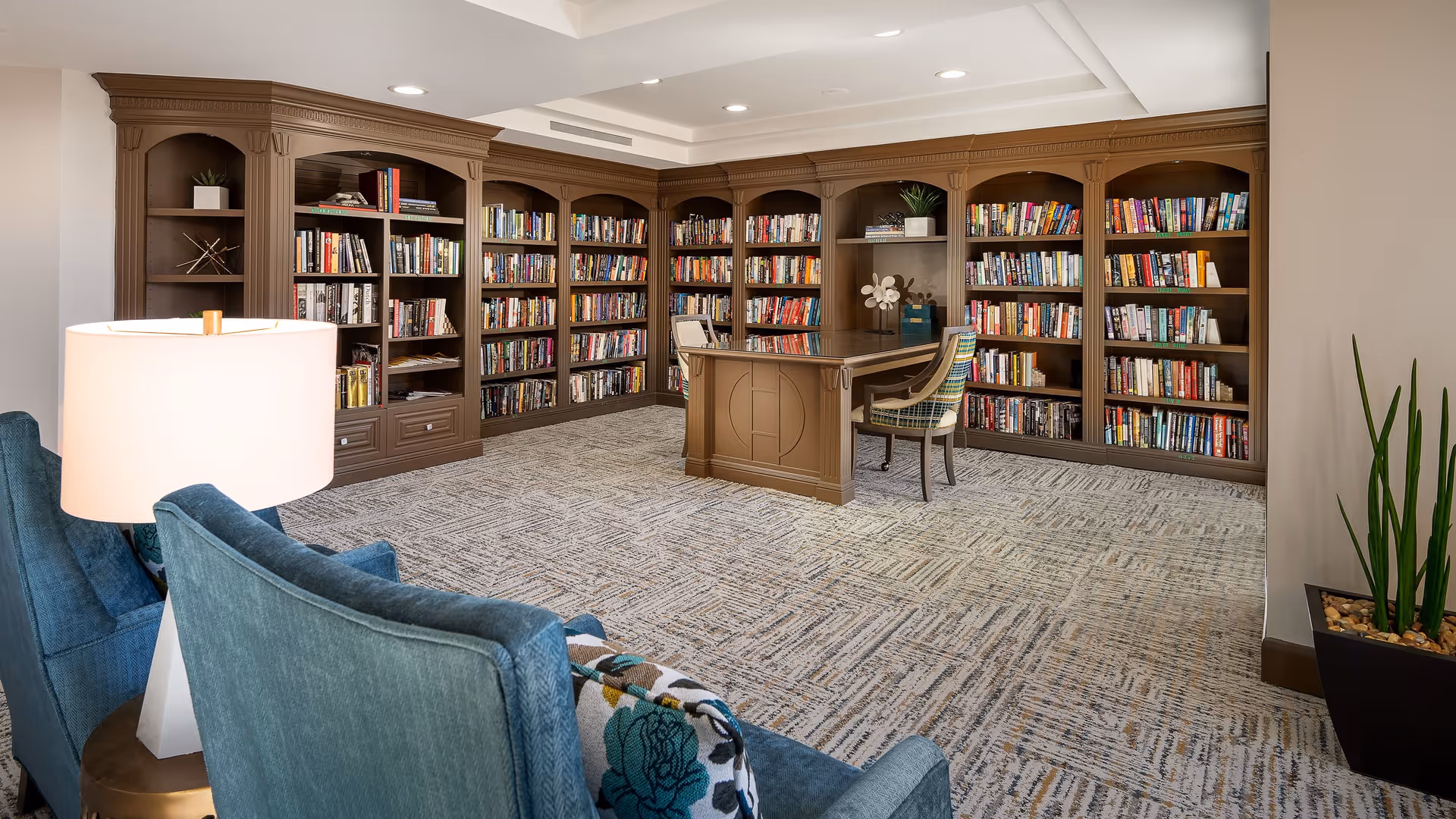A cozy library room with wooden bookshelves filled with books lining the walls. In the center, there is a wooden desk with a patterned chair. Two blue upholstered chairs with a floral cushion and a white table lamp are in the foreground. A tall plant in a black pot is placed near the right wall.