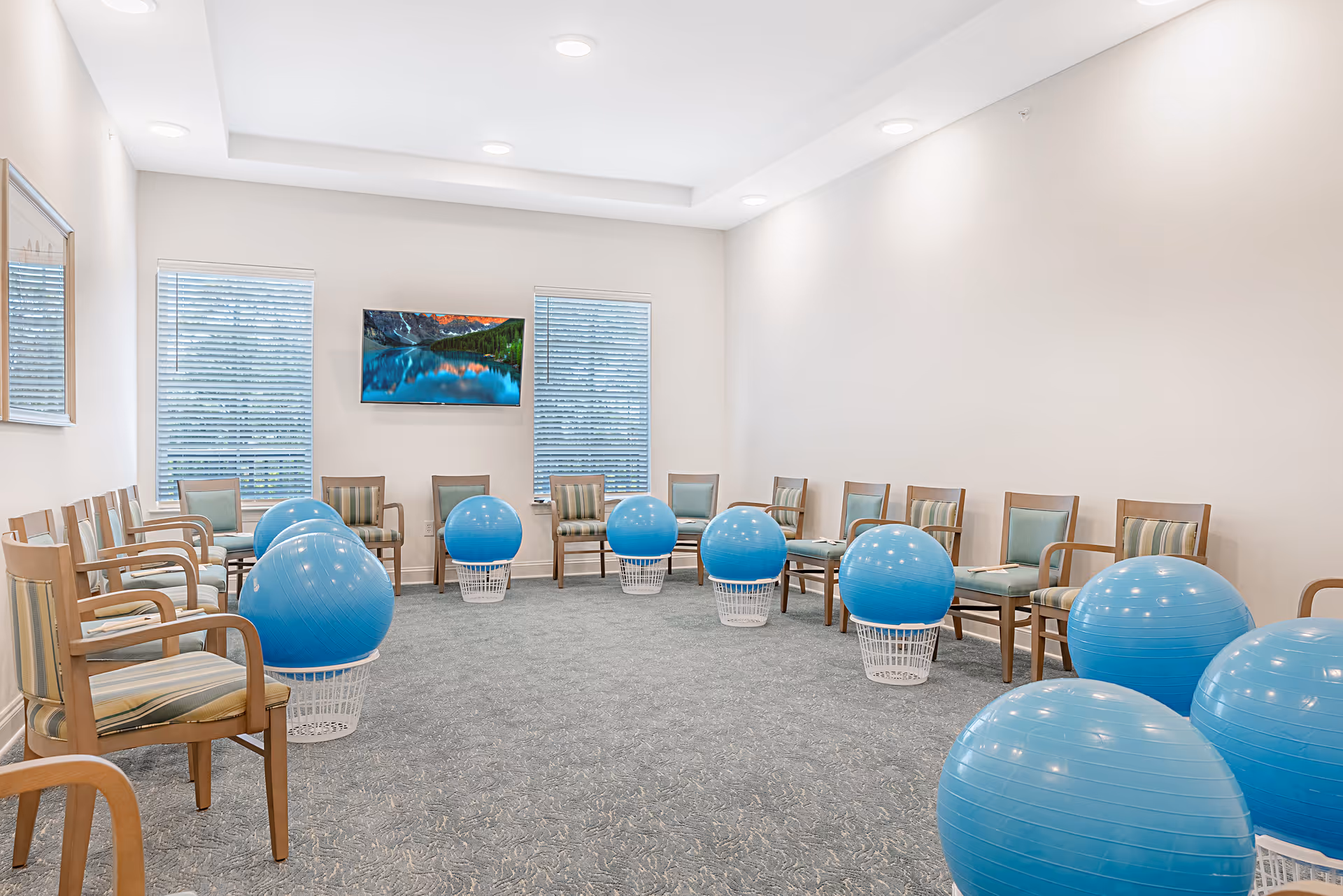 A spacious room with a circle of wooden chairs with striped cushions arranged around several large blue exercise balls placed on white baskets. The room has light-colored walls, two windows with blinds, a wall-mounted TV displaying a scenic lake and mountain view, and recessed ceiling lights.