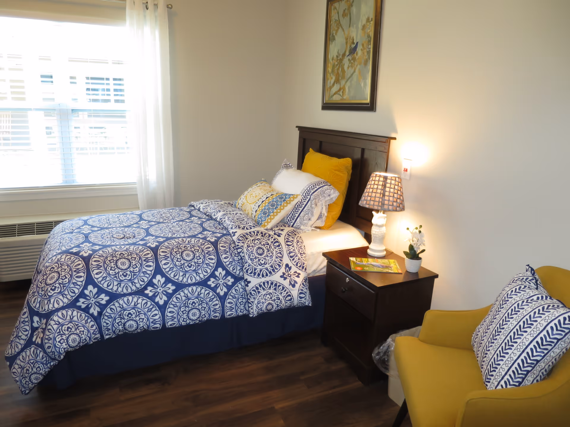 A cozy bedroom with a bed featuring blue and white patterned bedding and yellow accent pillows. Next to the bed is a dark wooden nightstand with a lamp, a small plant, and a magazine. A yellow armchair with a blue and white patterned pillow is positioned nearby. A window with white curtains allows natural light into the room, and a framed artwork hangs on the wall above the bed.